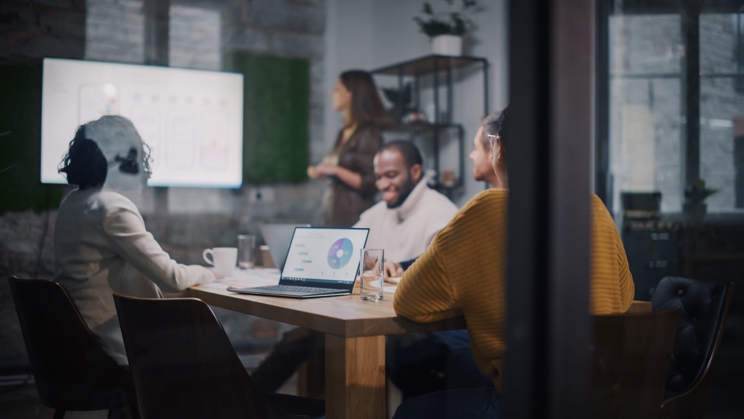 Team collaborating in modern office with laptops and presentation on screen, viewed through a glass partition.