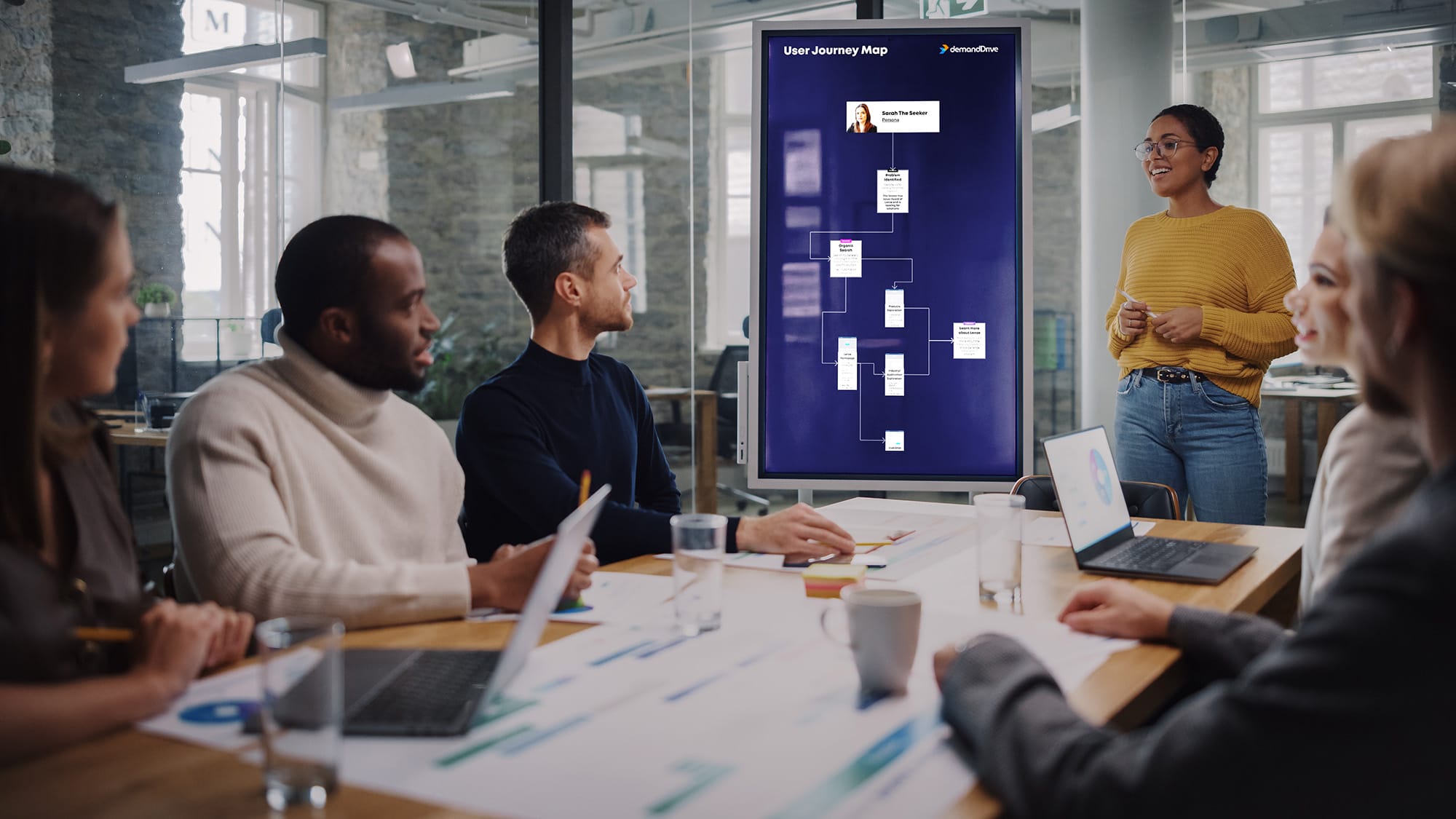 Team meeting with a woman presenting a user journey map on a screen, diverse group around a conference table.